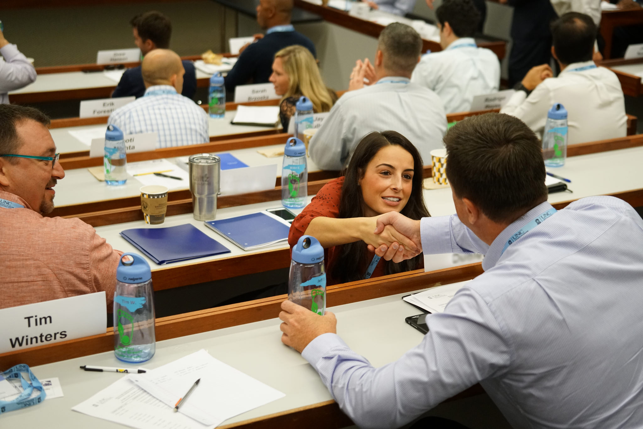 A man and woman in a classroom, reaching across a desk and shaking hands
