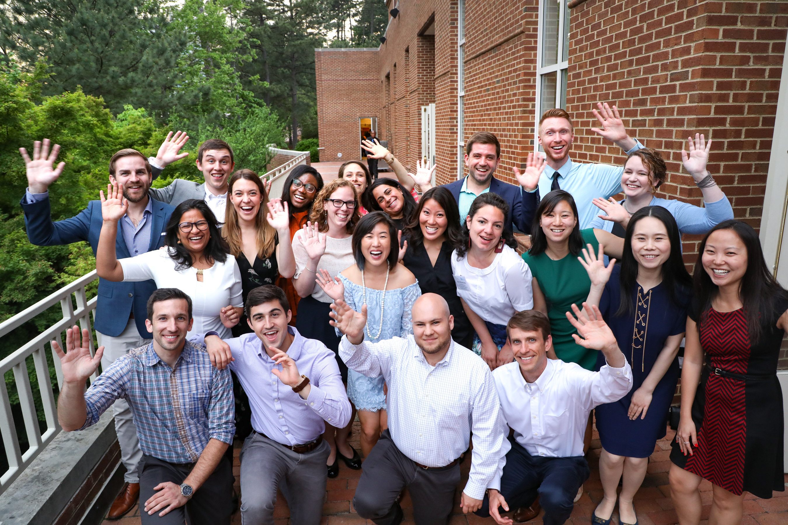Group of formally-dressed men and women outside of a building, smiling and waving at the camera