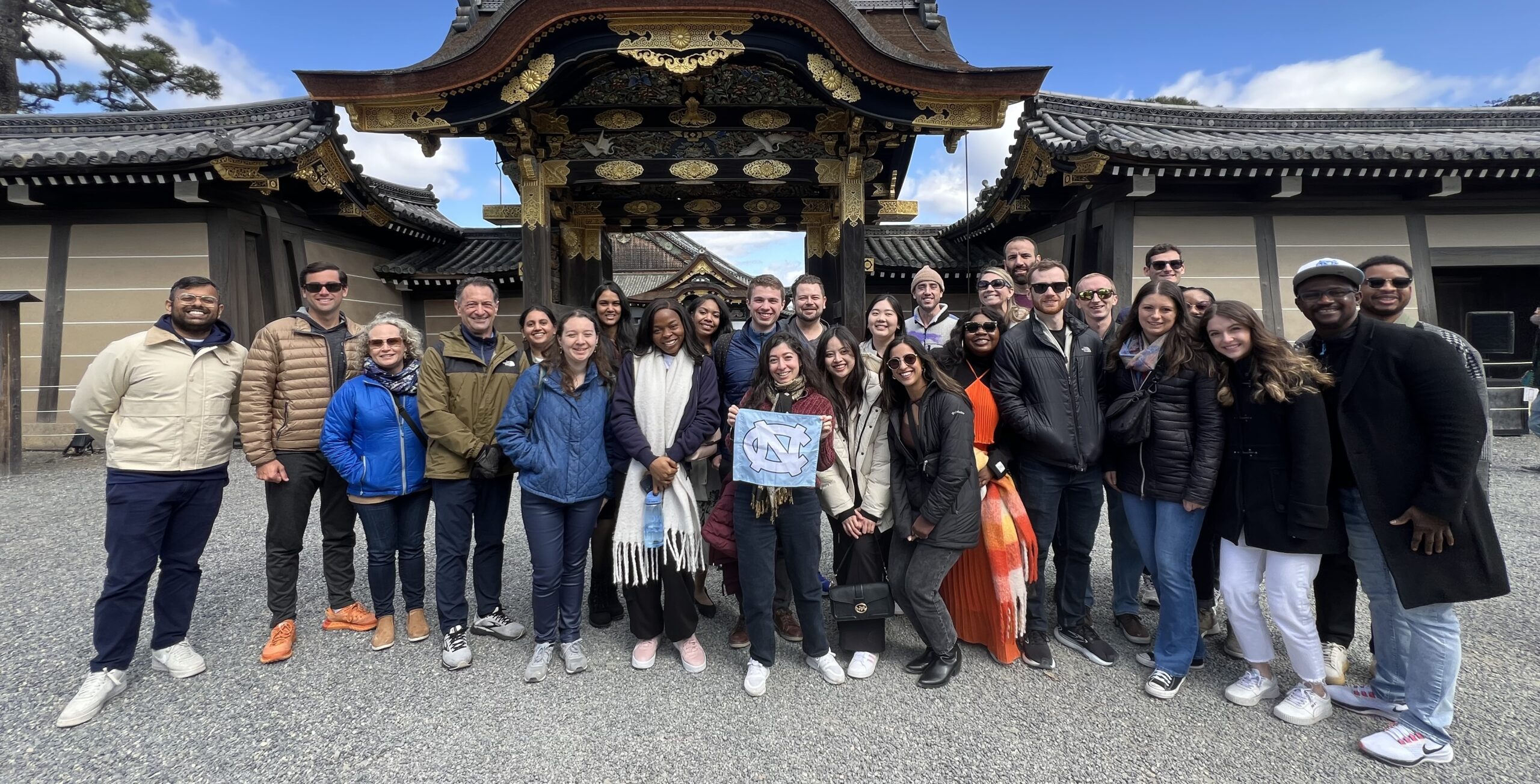 A group of students and faculty members in Thailand in front of ornate gate standing together and smiling at the camera holding a UNC flag