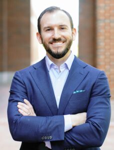 Matt Pirrello stands wearing a blue suit. He is smiling and has his arms crossed.