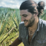 Alex Piasecki looks down and to the left, we can see his profile. He has long hair in a ponytail and is in some long grass.
