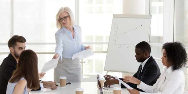 A businesswoman standing next to a whiteboard and handing reports to colleagues seated at a table.