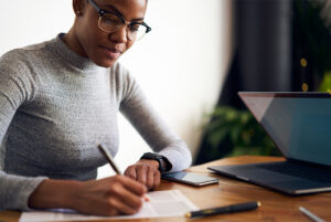 Businesswoman sitting at a desk writing on a piece of paper next to a laptop.