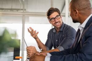 Two businessmen sitting at a desk talking.