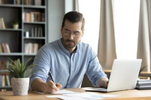 Businessman sitting at a desk in front of a laptop and writing on a piece of paper. 