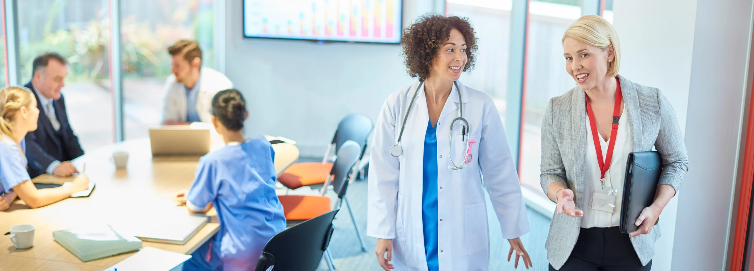 Two women walk through a doctors office. One is wearing business dress, while the other is in a white coat and stethoscope.