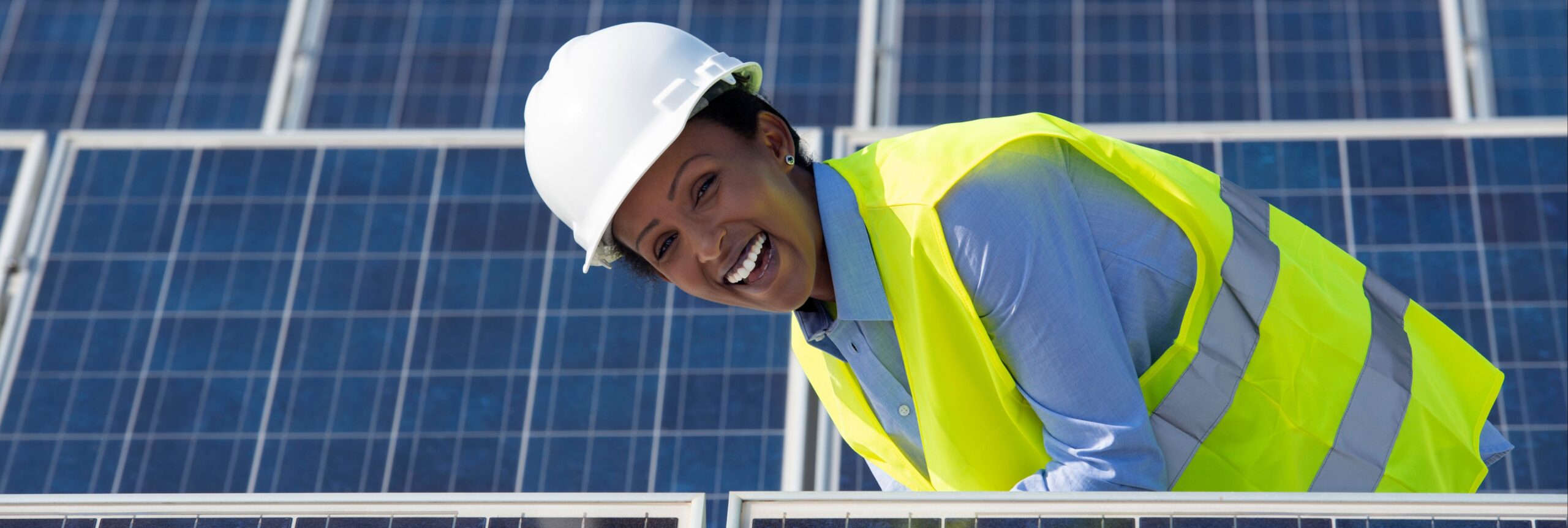 A woman stands in a solar farm smiling at the camera. She is wearing a yellow construction vest and hard hat
