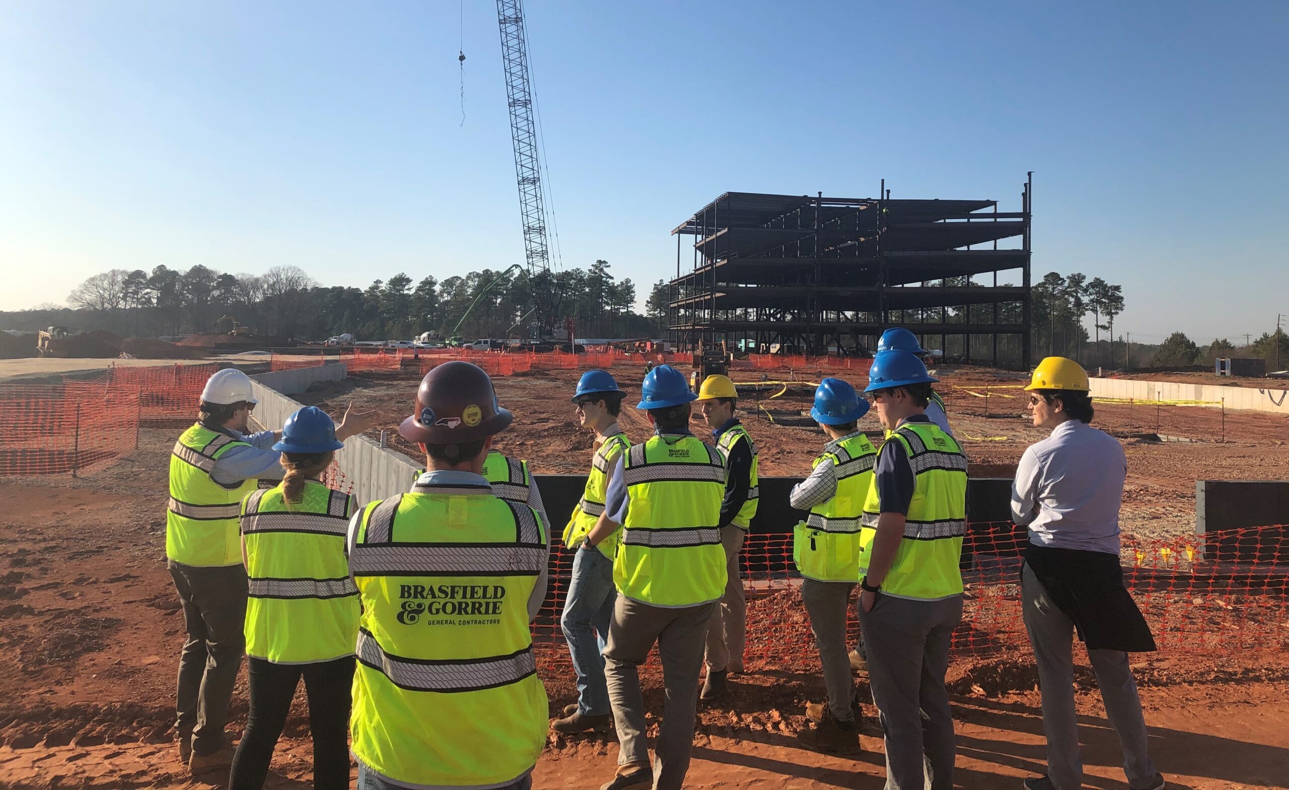 Approximately a dozen students observe a construction site from afar in hard hats and neon safety vests.
