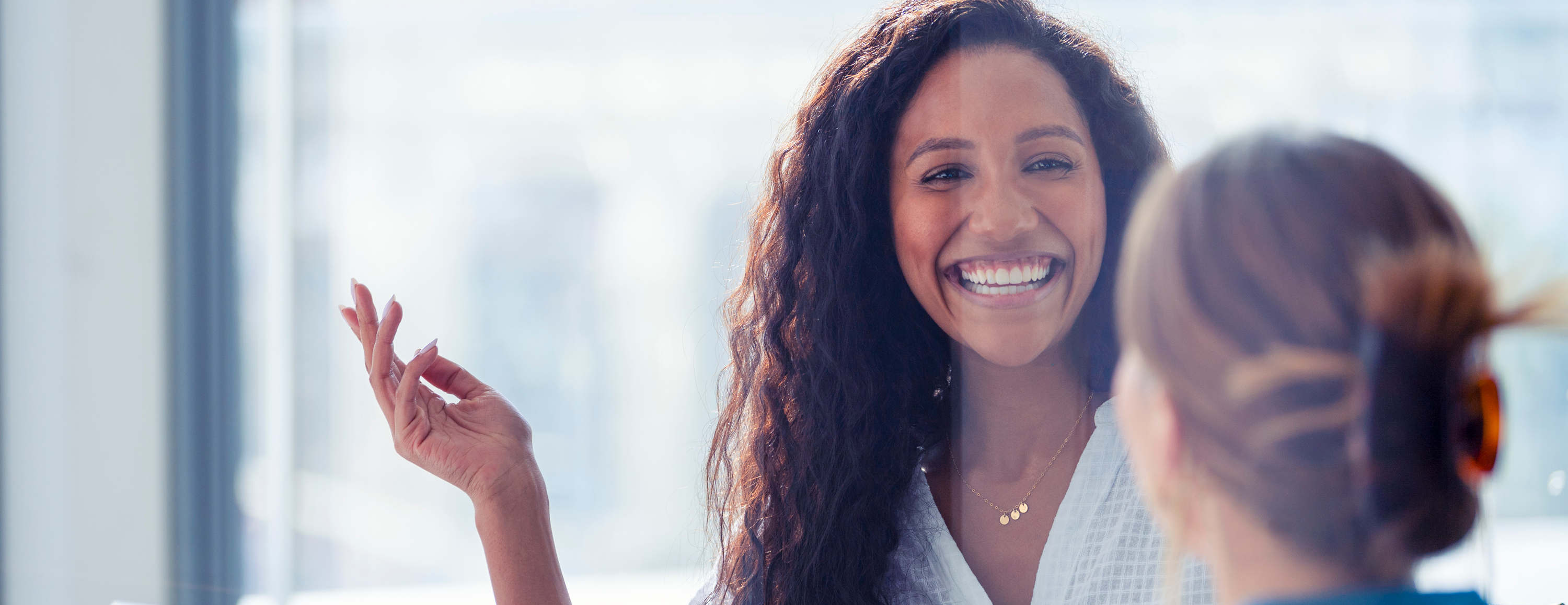 A woman is speaking to another person who is not facing the camera. The woman facing the camera is smiling a wide smile and gesturing with her hand as though she is talking.