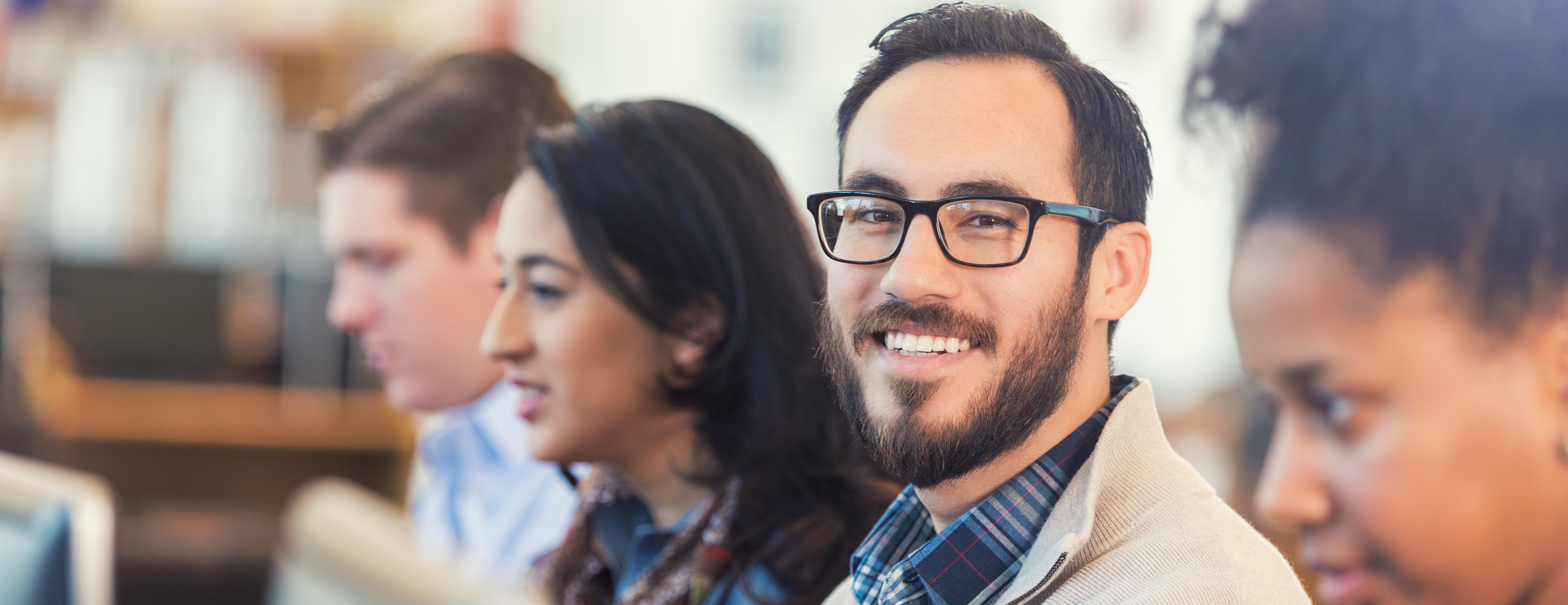 A student looks at the camera smiling. He is sitting next to other students who are focused on their work.