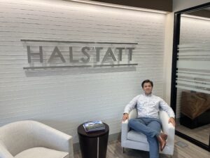 Aidan McConkey sits in a chair in the lobby of the Halstatt Real Estate Partners lobby. The Halstatt logo is on the wall behind him.