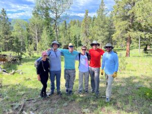 One woman and five men pose for a photo together in the Colorado wilderness surrounded but trees.
