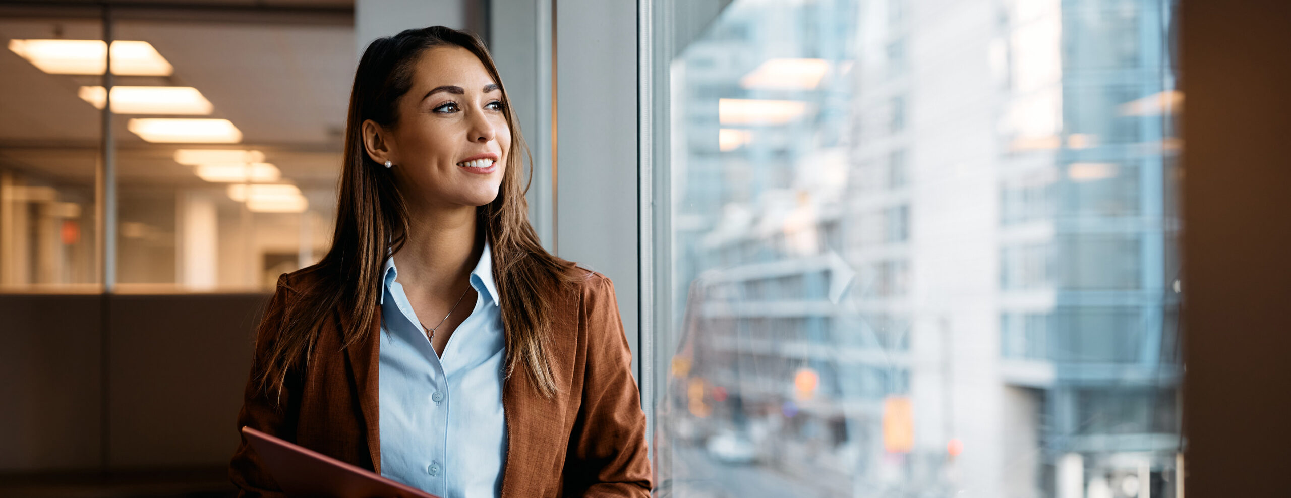 A woman in business casual clothing stands at a window looking outside. She is smiling slightly and seems optimistic and pensive.
