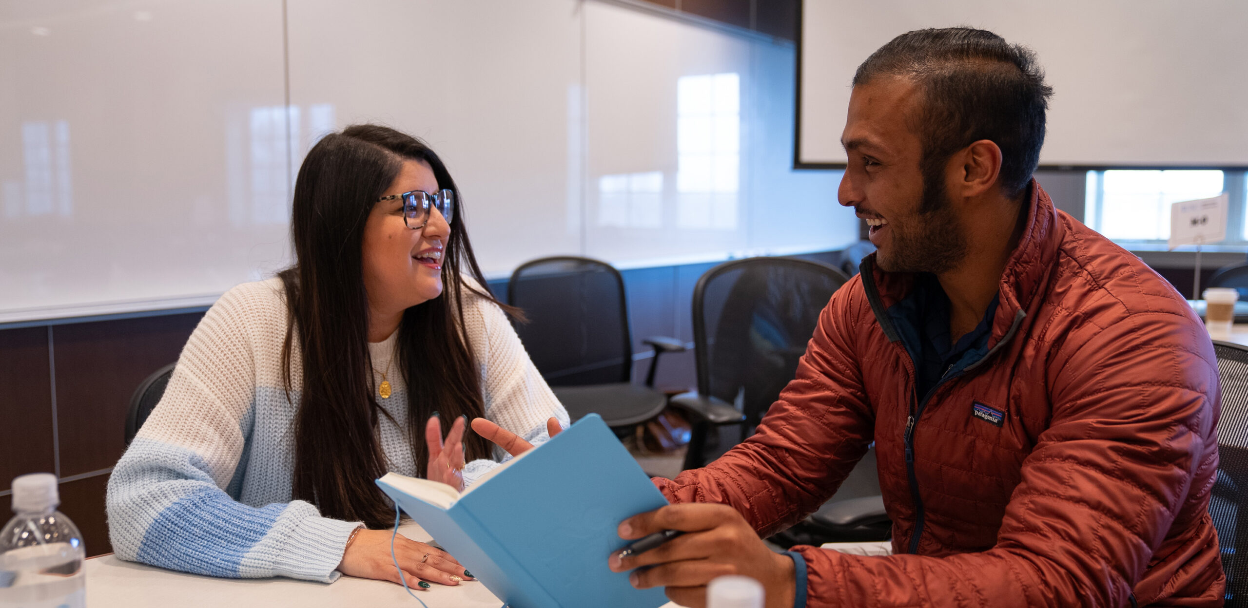 Two students are sitting at a table talking. One student is holing a notebook - it looks like they are collaborating on a project.