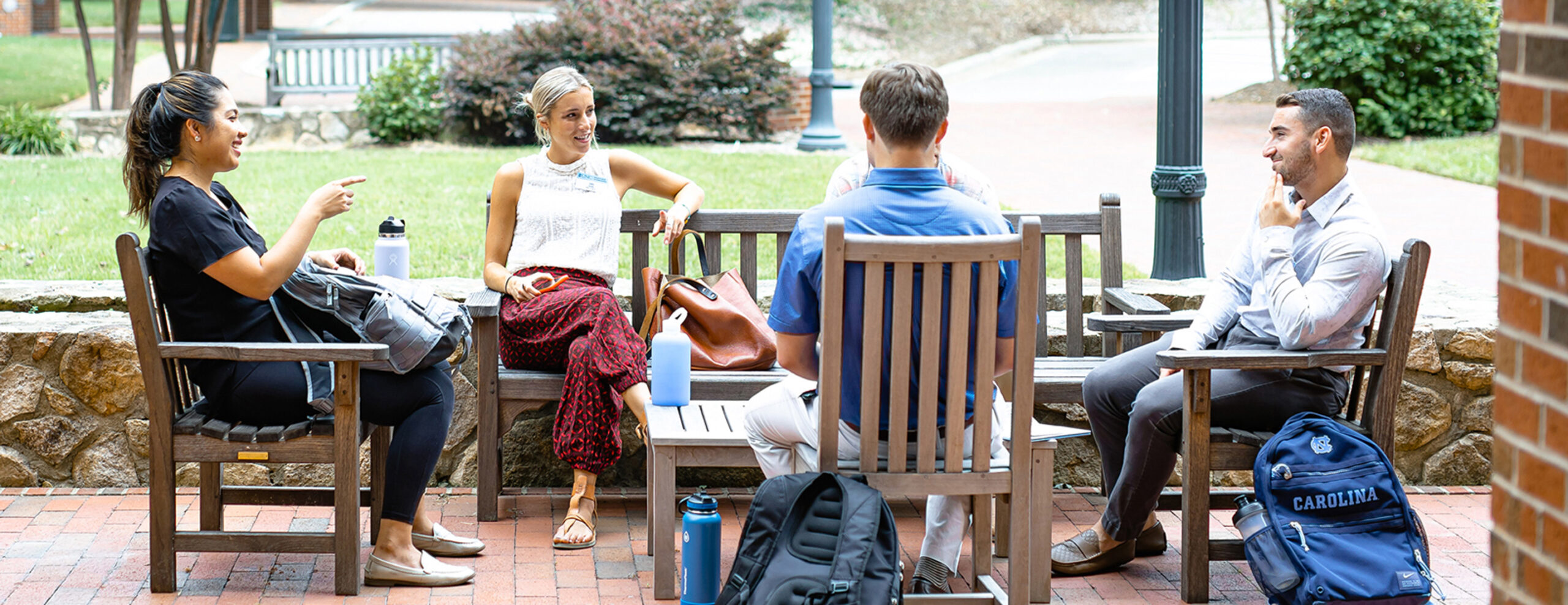 Four students are sitting outside in wooden chairs on a nice day. They are sitting casually and talking.