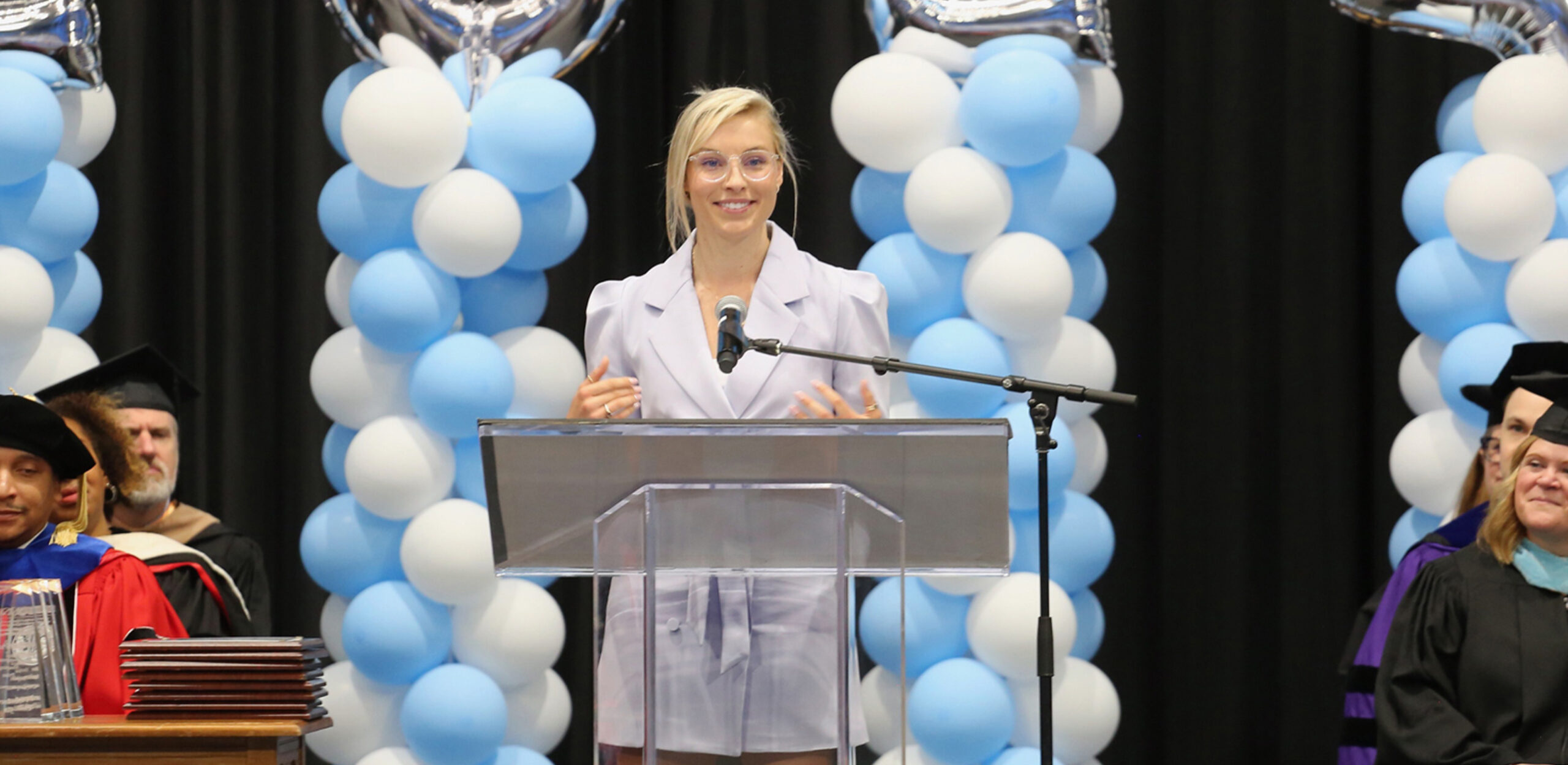 A woman stands at a podium. There are balloon towers behind her and people in graduation regalia sitting on either side, listening to her.