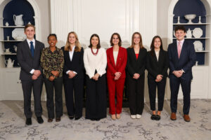 The Spring 2026 Phillips Ambassadors pose for a group photo in formal attire in front of a white and navy wall with displayed dishware.