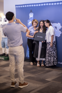 A MAC graduate poses with two others in front of a Carolina step and repeat as another person takes the photo.