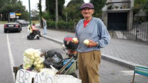Man in a hat holds two apples while leaning against his bike that holds bags of apples. He is selling them.