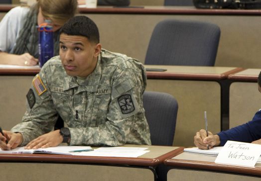Student in military uniform taking notes and looking to front of class, other students writing around him