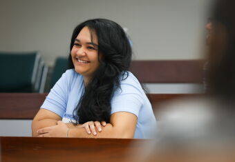 A woman sitting in a classroom leaning on her desk and smiling, facing slightly off camera.