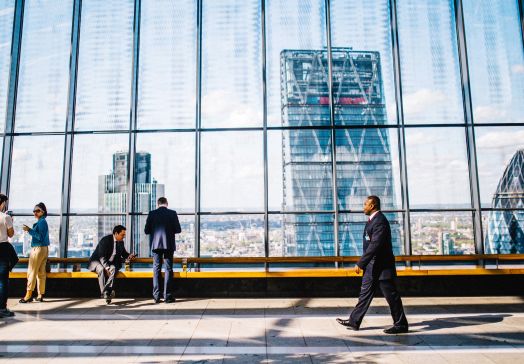 Man walking in a business suit in front of floor-to-ceiling windows revealing skyscrapers, with other people standing nearby
