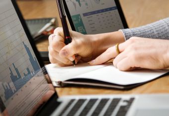 Person writing on a notebook, flanked by two laptops with graphs and maps