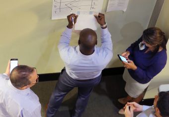 A group of men and women, two on their phones, surrounding a man referencing notes to add to a poster
