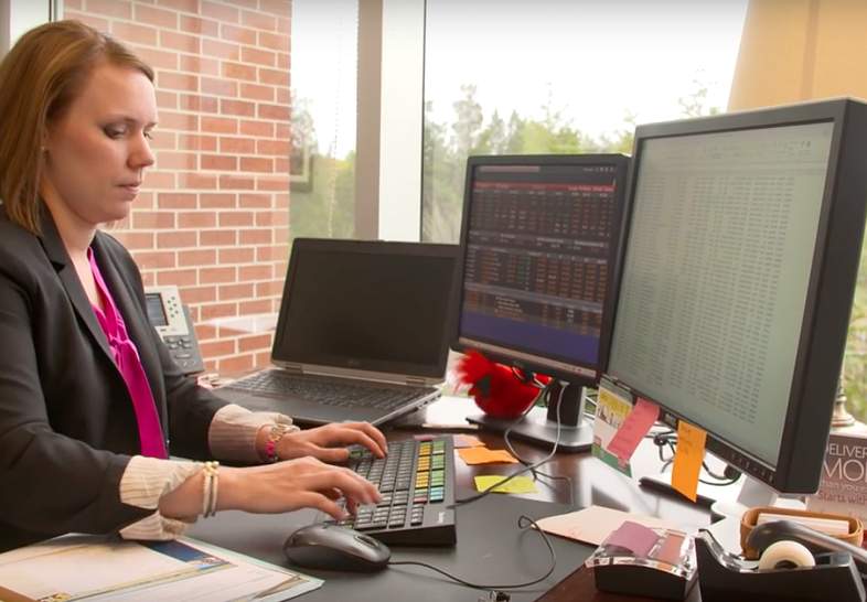 Leanne Fredericks facing two monitors and adding to a spreadsheet, flanked by notes and a laptop