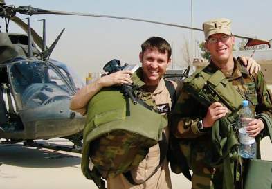 Brad Bertinot with another man, posing with military gear, in front of a military helicopter