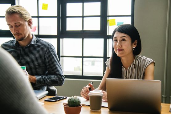 Woman sitting in front of a laptop at a table with a group, smiling and taking notes