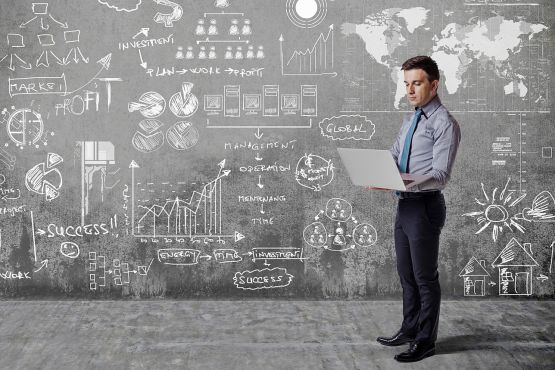 Man standing with a laptop in front of walls covered in business and financial terms and charts