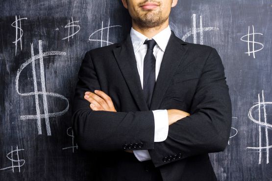 Man in suit with arms crossed standing in front of blackboard covered in dollar signs
