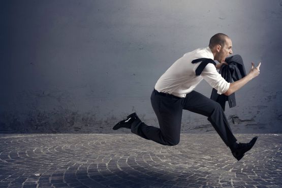 Man in suit carrying jacket, yelling as he leaps/runs over brick paved road in front of a gray wall