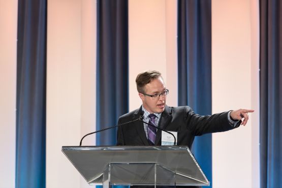 Scott Quilty speaking at a podium, with one hand pointing