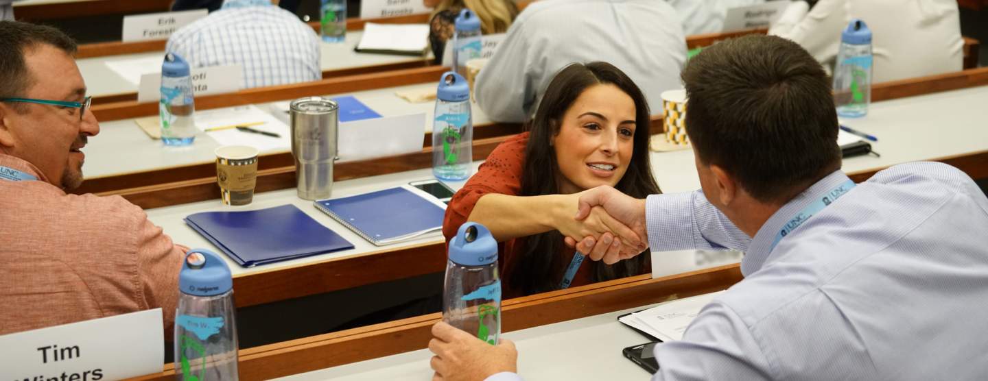 A man and woman in a classroom, reaching across a desk and shaking hands