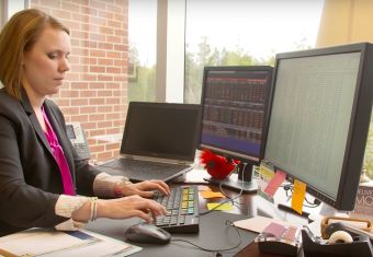 Woman facing two monitors and adding to a spreadsheet, flanked by notes and a laptop
