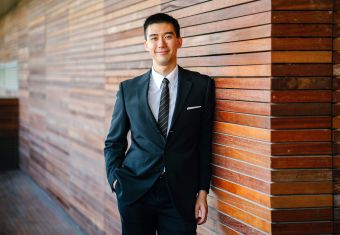 Man dressed in a suit, leaning casually against a wood-tiled wall
