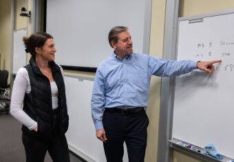Two people stand by a whiteboard, one is pointing at it while the other looks on and smiles.