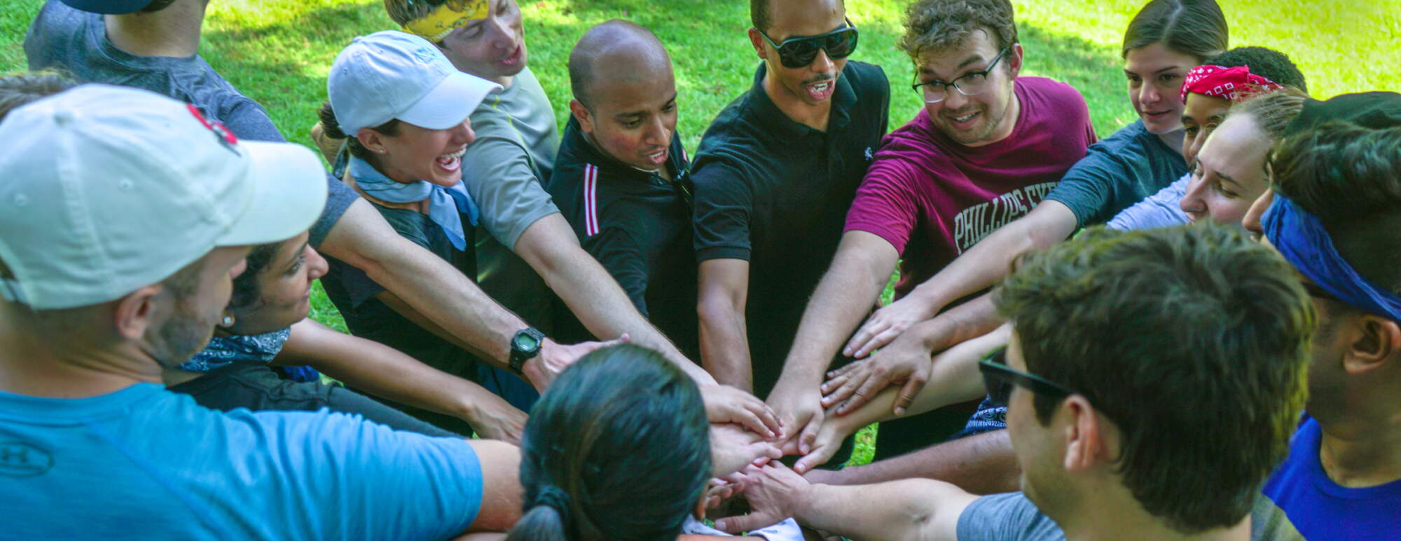 Group of casually-dressed men and women standing in a circle outside and stacking their hands on top of each other