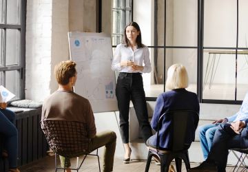 Businesswoman standing beside a whiteboard while giving a presentation to a group of seated businesspeople.
