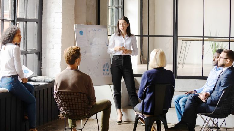 Businesswoman standing beside a whiteboard while giving a presentation to a group of seated businesspeople.
