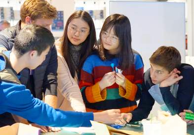 Standing around table six students animatedly discuss Makeathon project