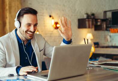 Businessman wearing headphones sitting in front of a laptop and waving into the webcam during a virtual meeting.