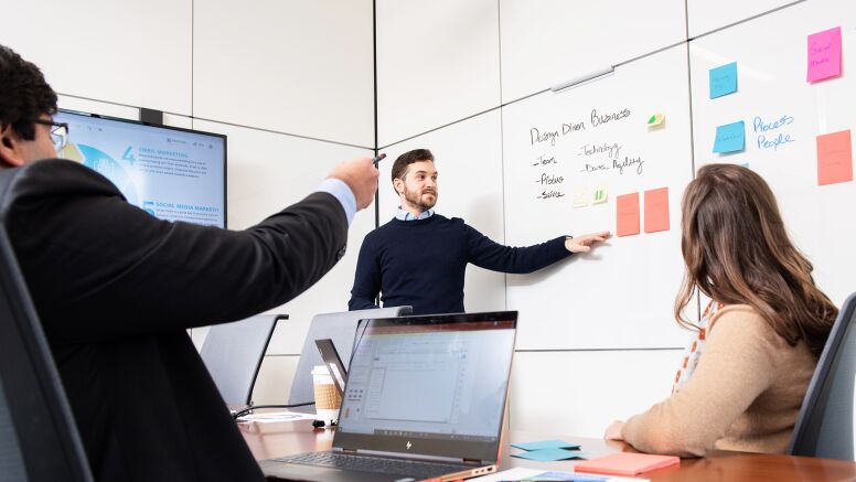 Three students around table discuss marketing terms written on whiteboard