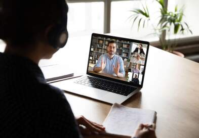 A businessperson in silhouette looking down at a laptop showing a presenter in a virtual meeting.