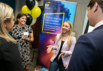 EY recruiter stands in front of branded sign and talks to MAC students at Structured Recruiting event.