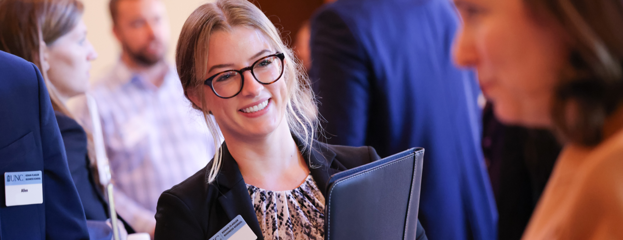 A woman with blonde hair and glasses looks off-screen, smiling. She is wearing business clothes.