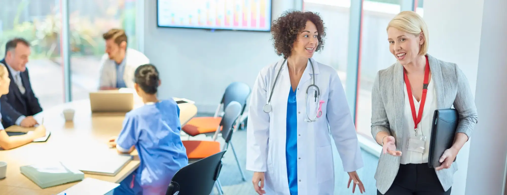 Two women walk through a doctors office. One is wearing business dress, while the other is in a white coat and stethoscope.