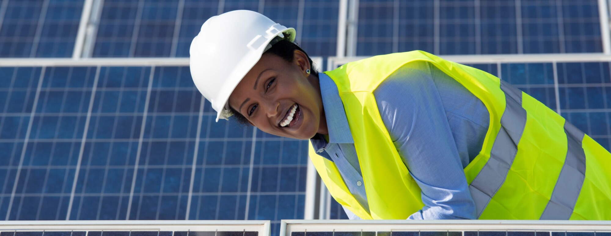 A woman stands in a solar farm smiling at the camera. She is wearing a yellow construction vest and hard hat